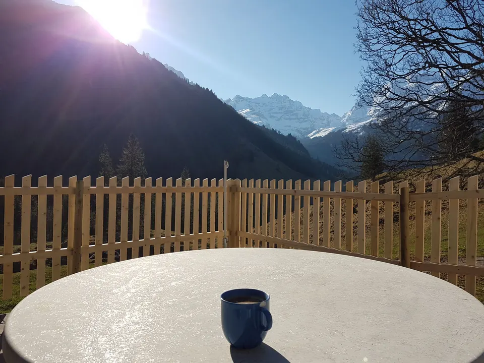 Tasse auf rundem Gartentisch, dahinter Holzzaun und verschneite Berge im Gegenlicht
