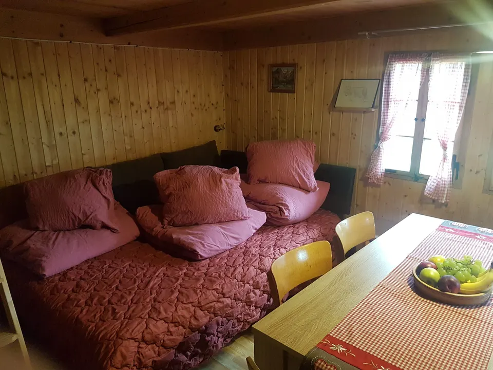 Breite Liegefläche mit roter Bettwäsche und Kissen in der Stube, daneben der Esstisch